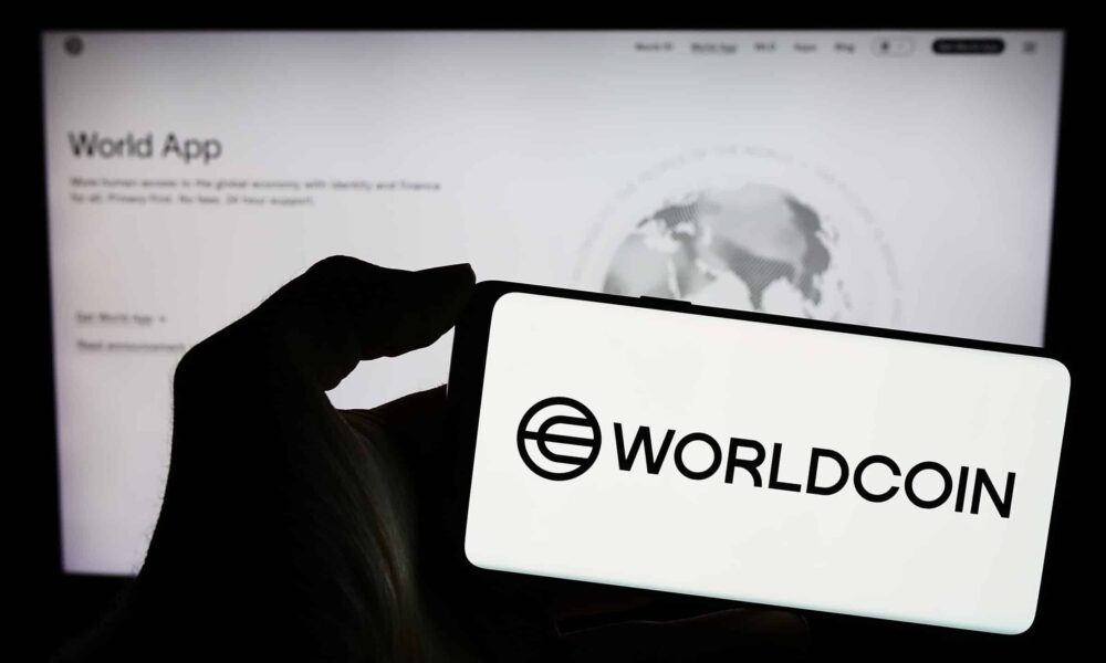 A photo of a hand holding a phone displaying "Worldcoin" and its logo. (Shutterstock/T. Schneider)